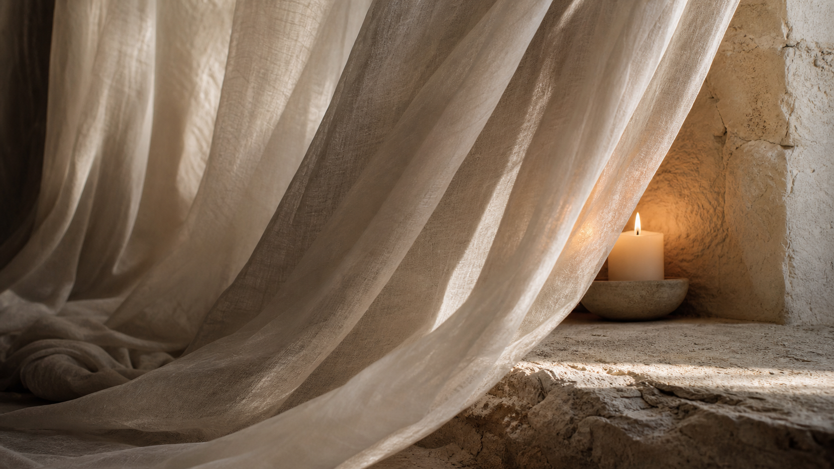 A sheer linen veil caught in soft light, partially revealing a stone wall and a glowing candle behind it — warm, muted tones